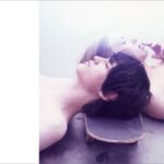 Two youth resting on a skateboard in soft lighting.