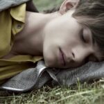Young man resting on grass, wearing a yellow shirt and striped jacket.