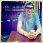 Young man in a cafe, wearing glasses and beanie, sitting at a table.
