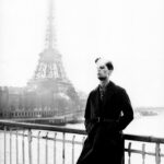 Man in trench coat by Seine River, Eiffel Tower in foggy backdrop.